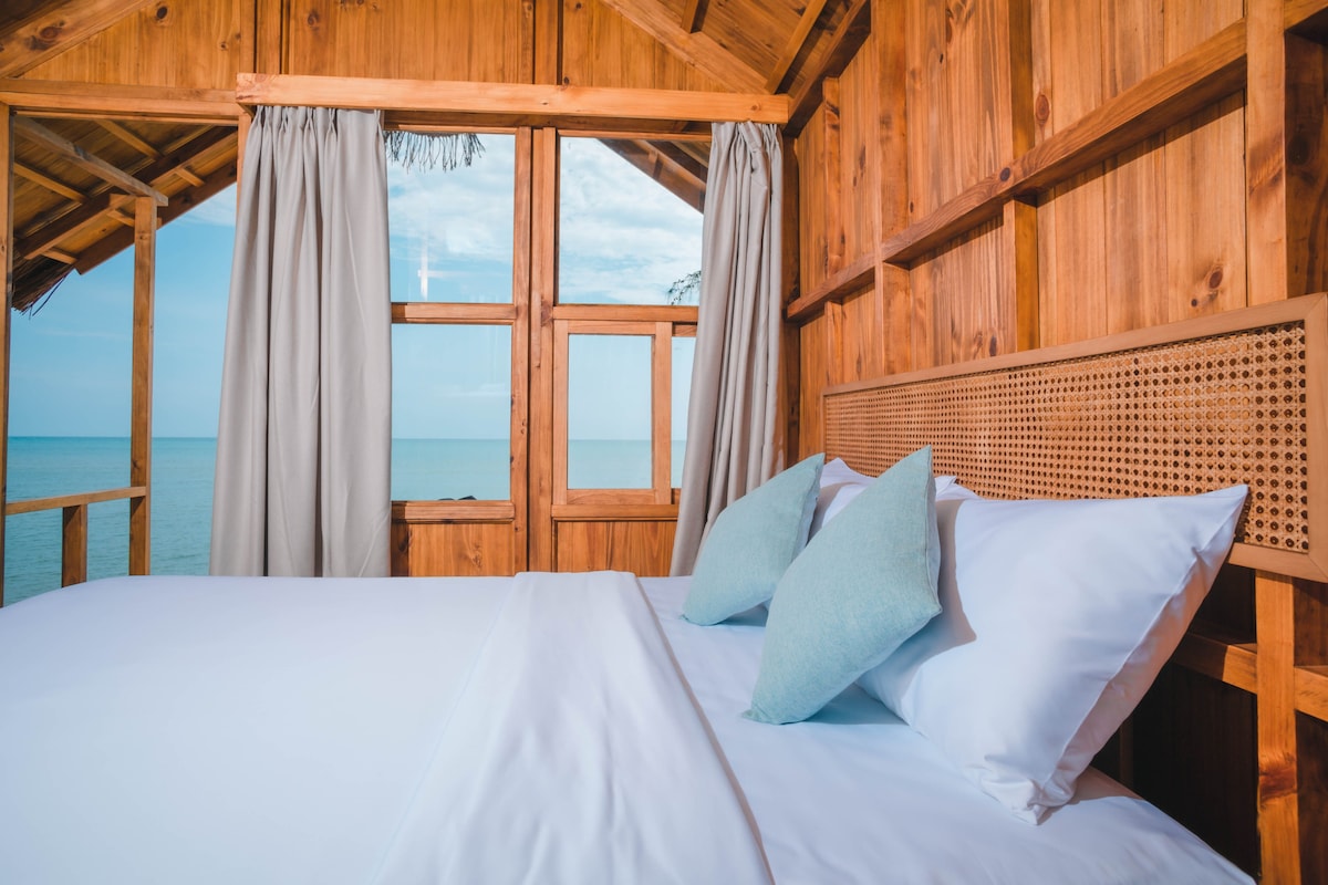A wooden bungalow interior features a spacious bed with crisp white linens and two aqua-colored pillows. Large windows frame a view of the calm sea, while beige curtains add softness to the natural wood walls.
