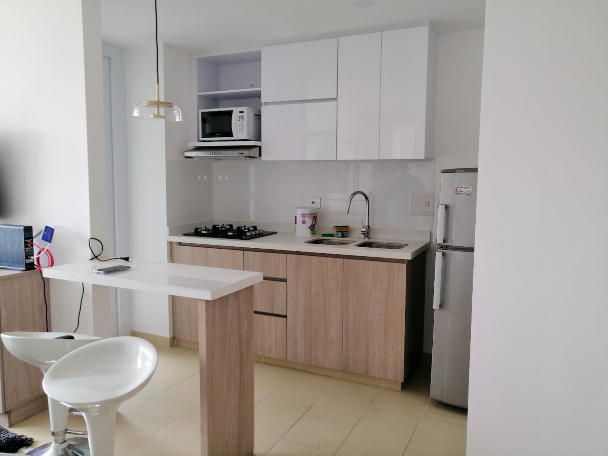 A modern kitchen area features light wood cabinetry and sleek white countertops. Essential appliances, including a refrigerator and a microwave, are included. A gas stove and sink are situated on the counter, while a small breakfast bar with two stools provides additional seating.