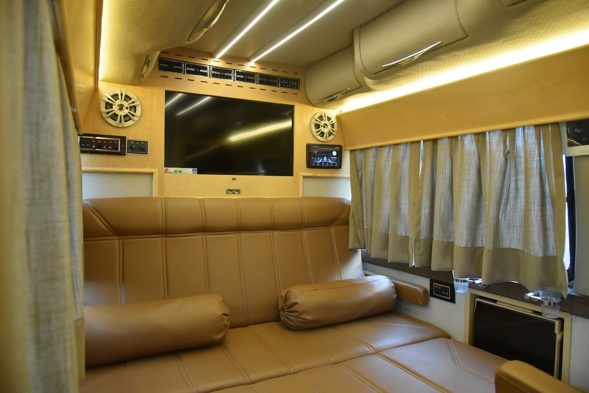The interior of the campervan features a spacious seating area with a brown leather sofa, which includes two backrest cushions. Above, a flat-screen LED display is mounted, complemented by sound systems on both sides. Soft, illuminated lighting enhances the warm environment.
