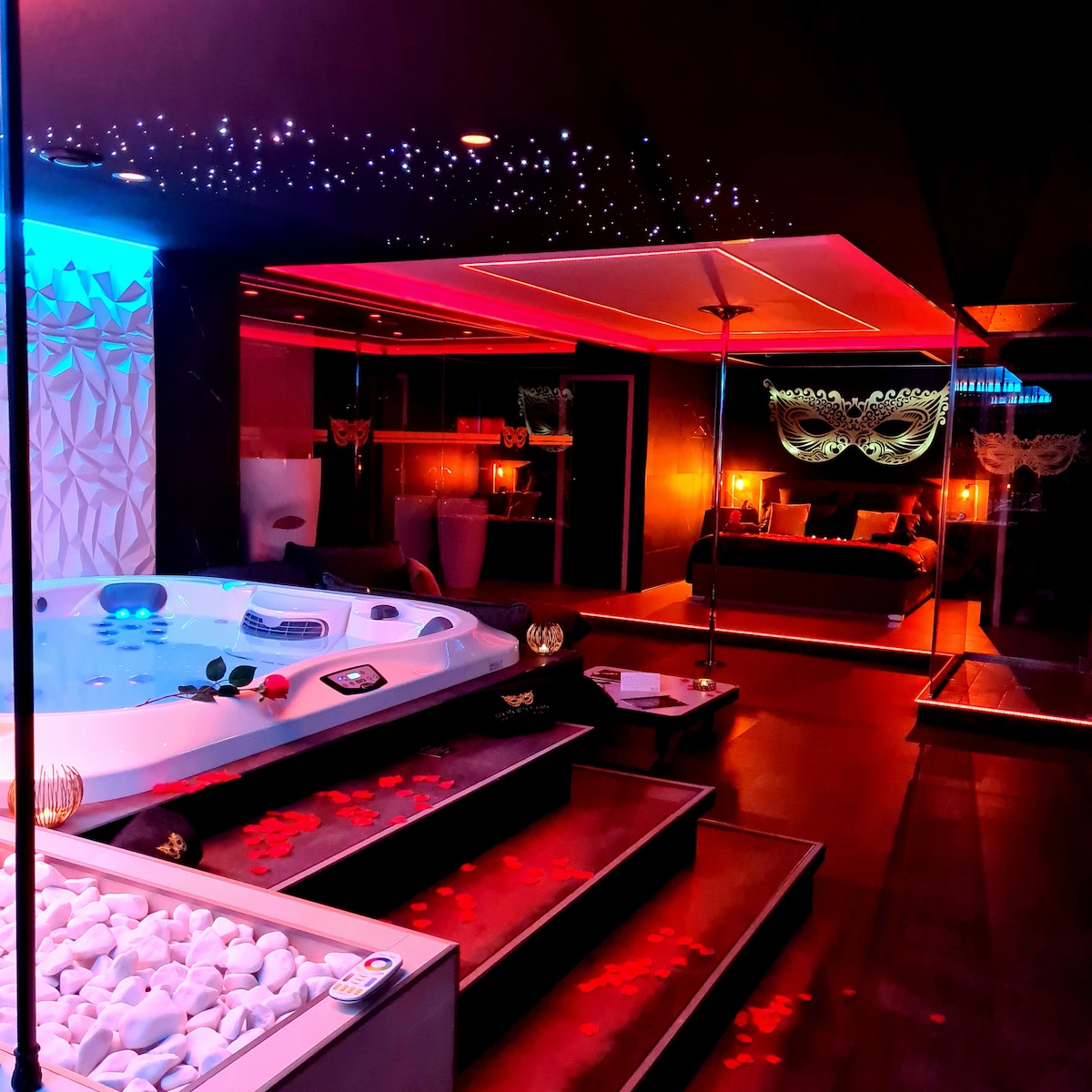 A modern suite is illuminated in soft hues of blue and red, featuring a luxurious jacuzzi with rose petals scattered around. A spacious bed is visible behind glass partitions, accented by ambient lighting and decorative elements, including a starlit ceiling above.