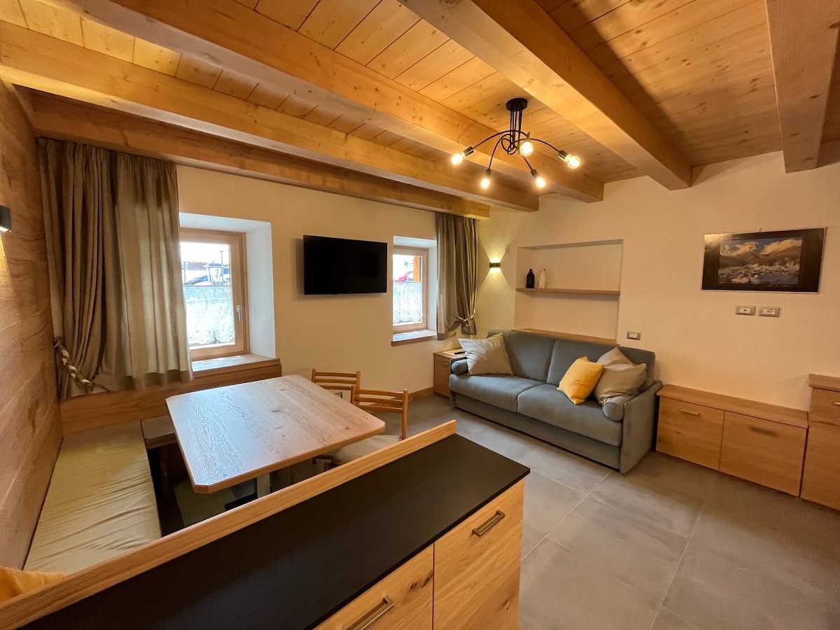 The living area features wooden beam ceilings and large windows, providing natural light. A cozy sofa is paired with decorative cushions. A dining table is positioned nearby, complemented by minimalist chairs. The walls are adorned with subtle decor, enhancing the room's inviting feel.