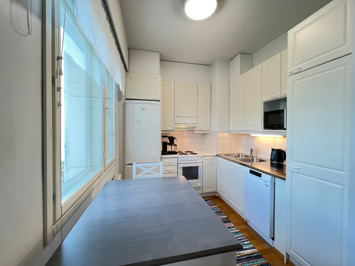 A bright kitchen features white cabinetry and modern appliances. A large window allows natural light to fill the space, while a wooden countertop extends for meal preparation. A patterned rug adds a touch of color, complementing the overall clean and functional layout.