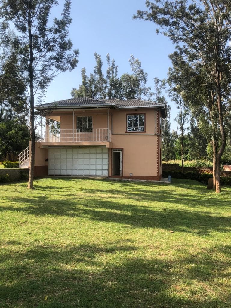 A two-story house is set amid a verdant lawn, surrounded by tall trees. A spacious balcony overlooks the yard, and a large garage door is visible at the ground level. Natural light brightens the exterior, creating a serene setting.