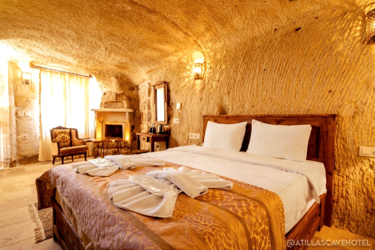 A cave room showcases a spacious bed with neutral linens and an ornate blanket. A fireplace is positioned near the foot of the bed, and natural light filters through the window over sheer curtains. A small seating area with a chair is visible in the corner.