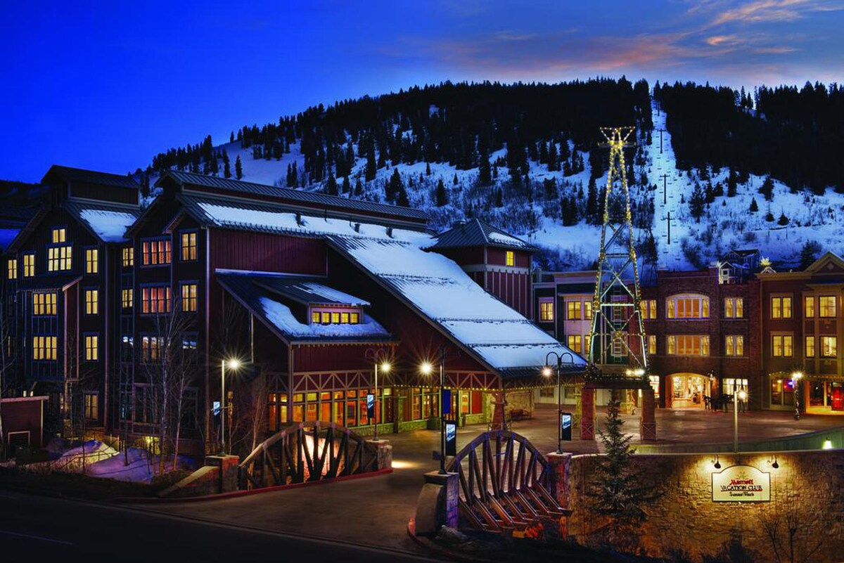 The exterior of the building is shown in the evening, with a snow-covered roof and warm lights illuminating the windows. A festive tree and decorative lights are visible, with a mountain backdrop featuring skiing trails.