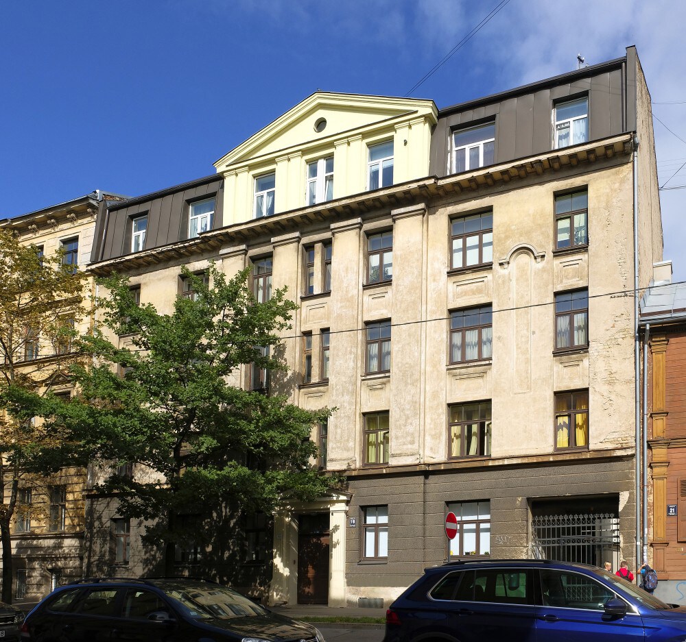 The exterior of a vintage-style building is depicted, showcasing a blend of light yellow and muted beige colors. Large windows are visible on multiple levels, framed by dark awnings. Nearby, a tree provides greenery, and parked cars line the street.