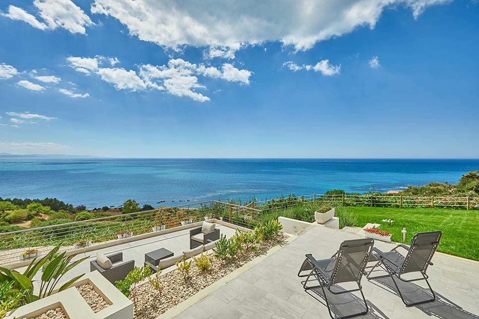 A spacious outdoor terrace is visible, featuring comfortable lounge seating and two reclining chairs. The terrace overlooks a panoramic view of the serene blue sea and sky, with greenery and flowers adding a touch of nature to the surrounding landscape.