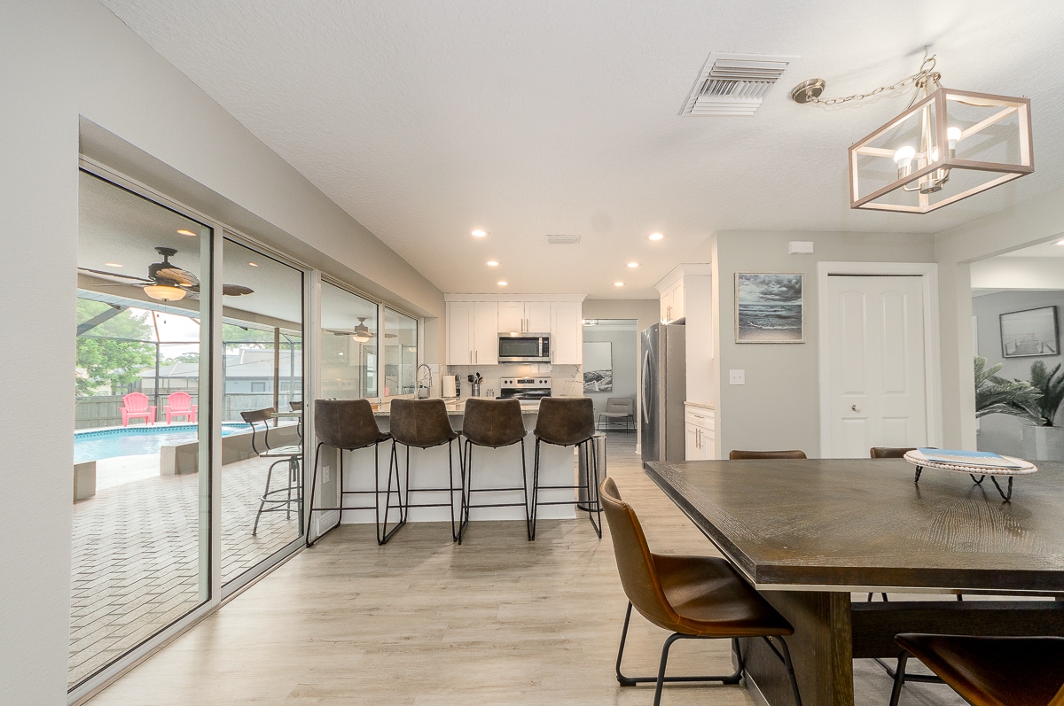 The open-concept kitchen and dining area feature modern furnishings. A large table provides seating for six, while a breakfast bar with four cushioned stools offers additional dining options. Natural light flows through expansive glass doors, creating a seamless connection to the outdoor pool area.