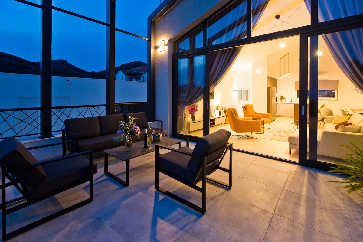An outdoor terrace is highlighted by a modern seating arrangement consisting of four cushioned chairs and a coffee table. Soft lighting illuminates the area, with sheer curtains framing expansive glass doors that lead to the indoor living space.