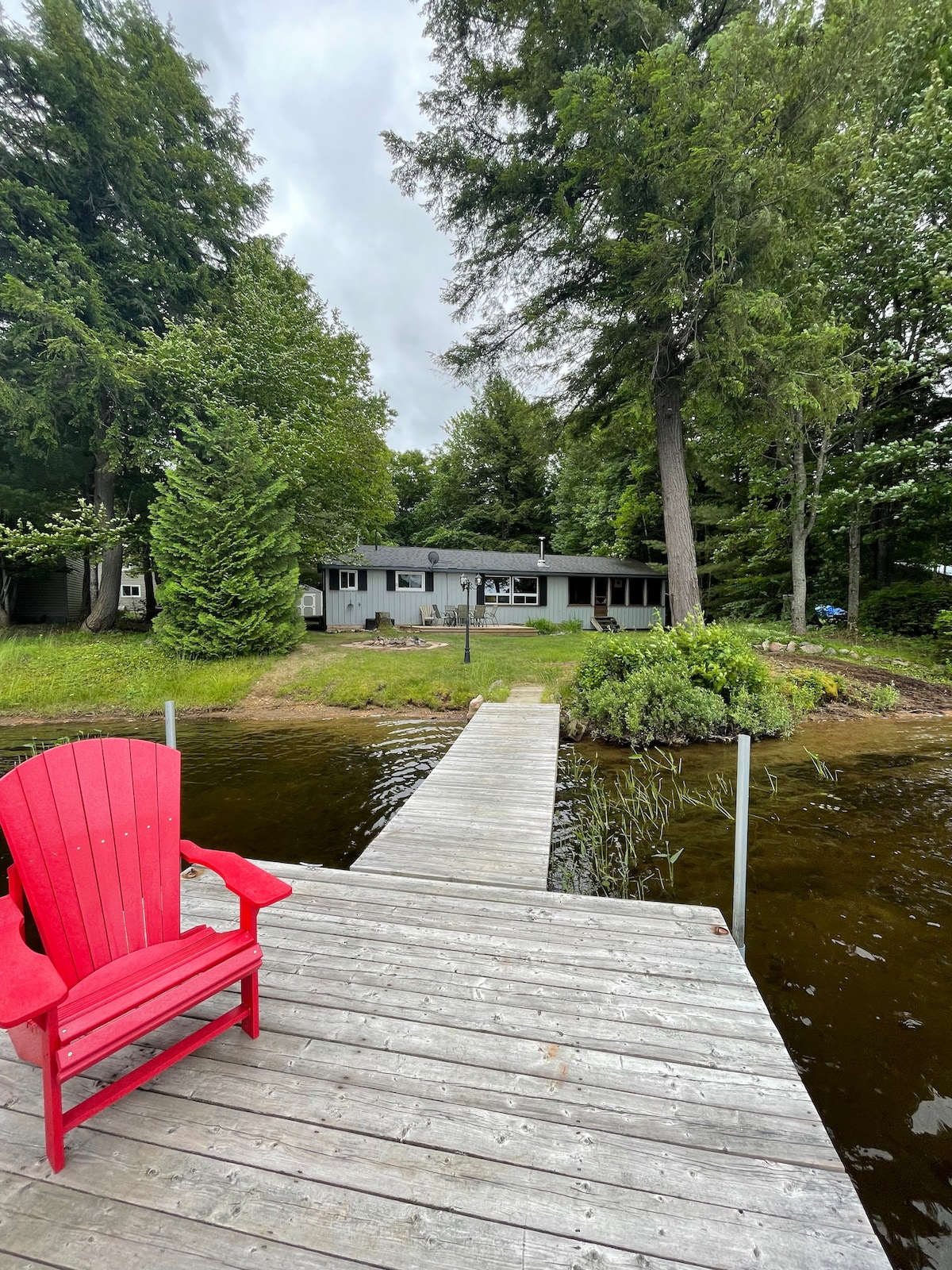 A wooden dock extends into the calm waters of Wren Lake, with a vibrant red chair placed at the front. In the background, the cottage is surrounded by lush trees, creating a serene atmosphere. The sky is overcast, adding to the tranquil setting.