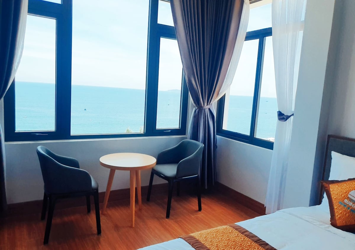 A bright room features large windows that offer a panoramic view of the ocean. Two dark chairs are positioned beside a round table, creating a cozy seating area. Soft curtains frame the windows, allowing natural light to fill the space.