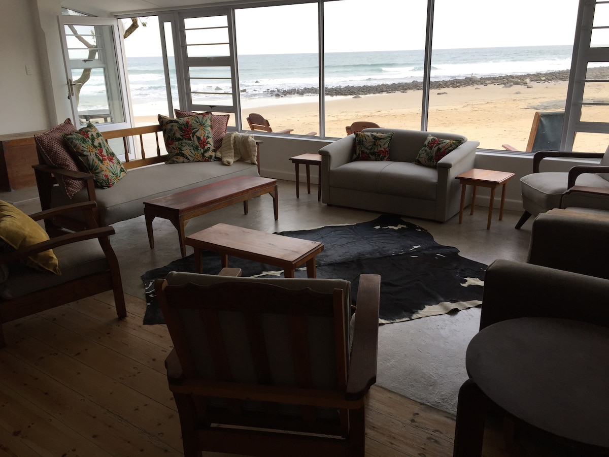 A bright living area features large windows offering views of the beach and ocean. Comfortable seating includes a mix of armchairs and sofas adorned with colorful cushions. A wooden coffee table is centrally placed on a rug, with a side table providing additional surface space.