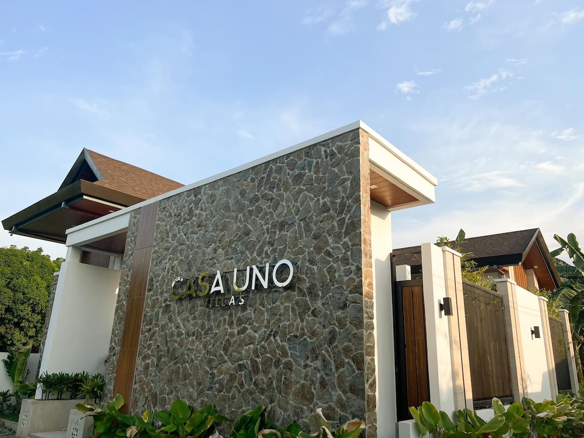 The image displays the exterior of Casa Uno Villas, featuring a modern stone façade with the property's name prominently displayed. Green foliage lines the entrance, and the structure is complemented by architectural elements like a sloped roof and wooden accents, all set against a partly cloudy sky.