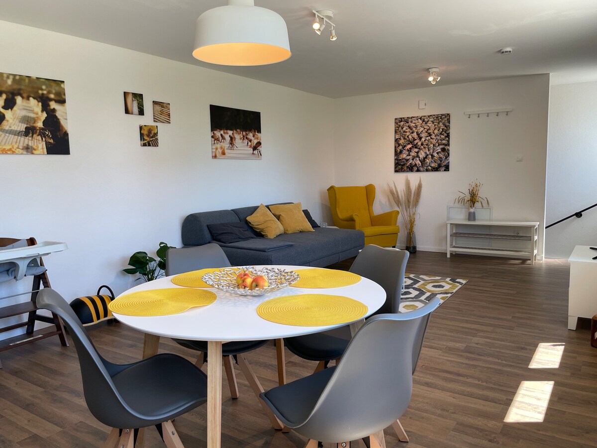 A bright living and dining area is featured, showcasing a modern round table surrounded by sleek chairs. A cozy sofa is accented with cushions, and vibrant wall art adorns the walls. Natural light floods the space, highlighting a warm and inviting ambiance.