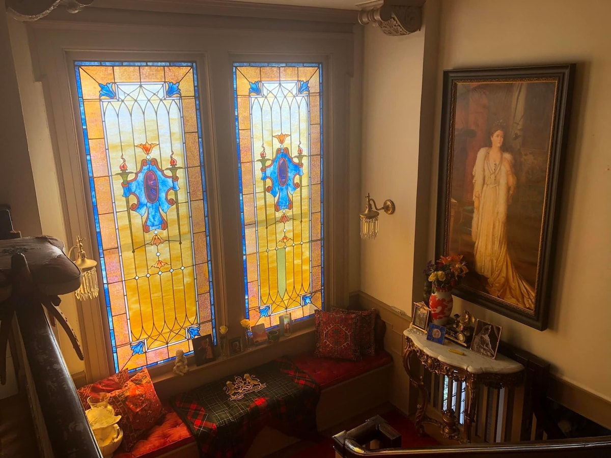 A stairwell nook features two large stained glass windows, casting colorful light into the space. A comfortable seating area with decorative cushions is positioned beneath the windows. A framed painting hangs on the wall nearby, accompanied by a small table adorned with photographs and flowers.
