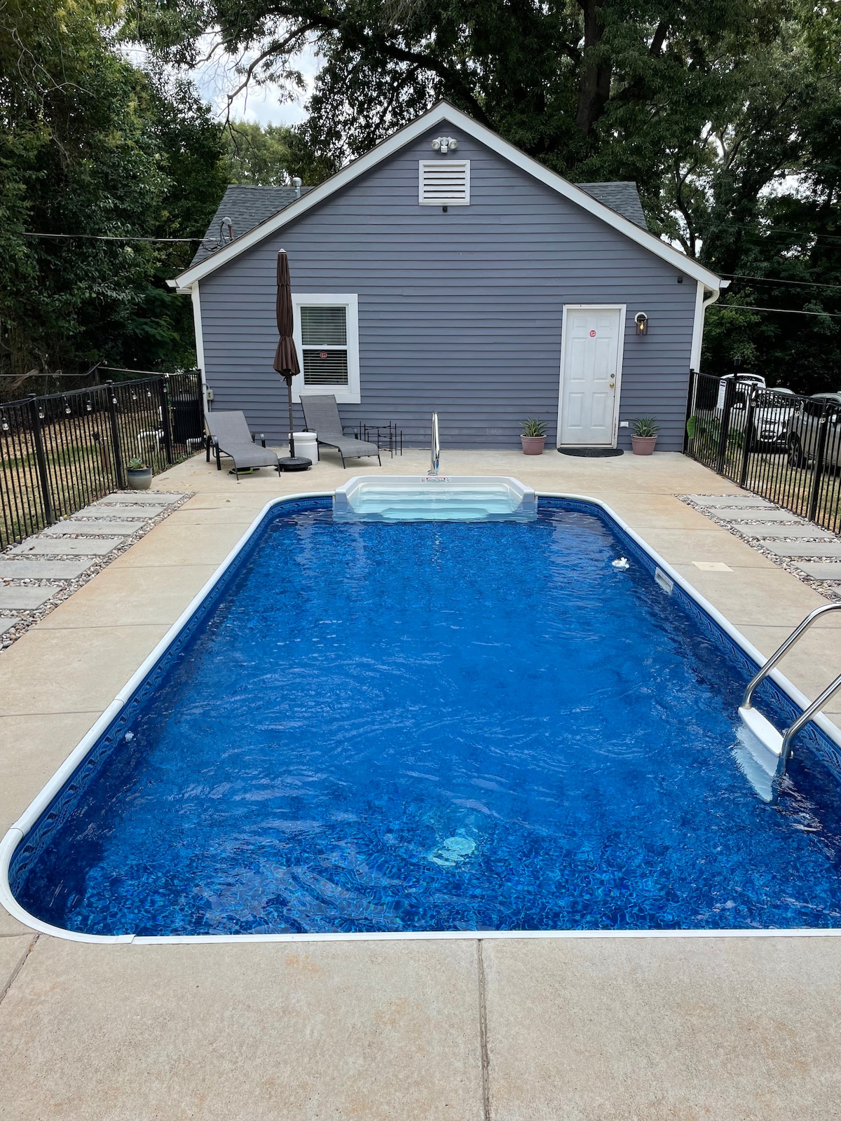 A private pool is surrounded by a paved patio, featuring lounge chairs and an umbrella for shade. The pool area is enclosed by a black fence, with steps leading into the clear water. The bungalow's exterior is visible in the background.
