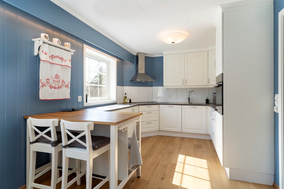 A well-equipped kitchen features white cabinetry and a spacious wooden island with seating for two. A modern hood and stovetop are positioned against a blue wall, while a window allows natural light to illuminate the area. Neutral tones and a tiled backsplash contribute to the clean design.