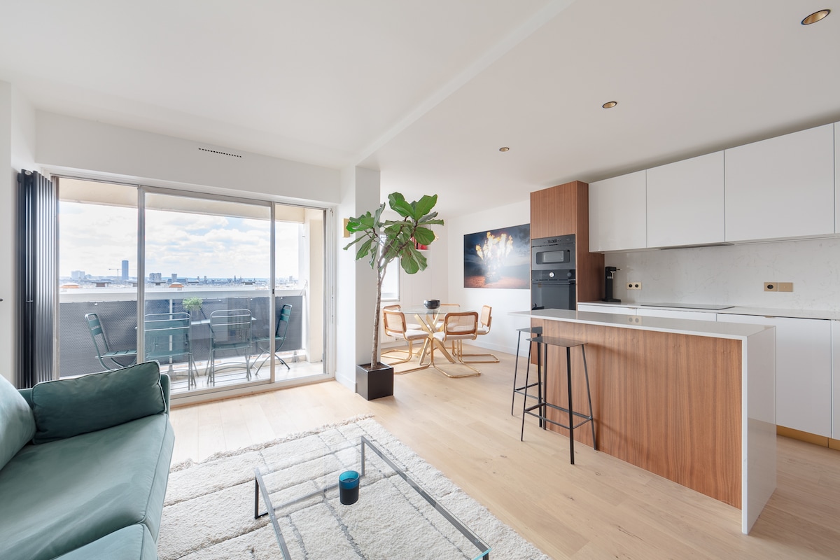 The open-plan living space features an inviting sofa and modern kitchen with wood accents. A large window fills the room with natural light and offers a view of the balcony with outdoor seating. A potted plant adds a touch of greenery to the atmosphere.