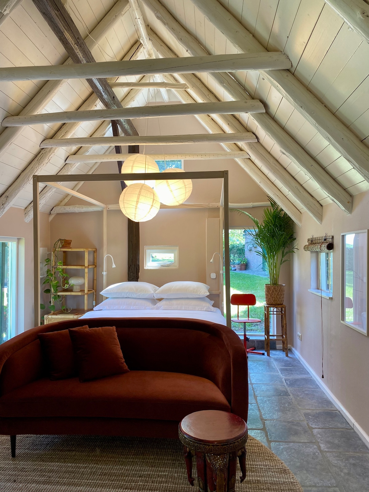 An airy interior features a spacious bed framed by a minimalist canopy, beneath a sloped ceiling with exposed beams. A cozy seating area is provided with a deep red couch and a round side table. Natural light streams in through large windows, enhancing the tranquil atmosphere.