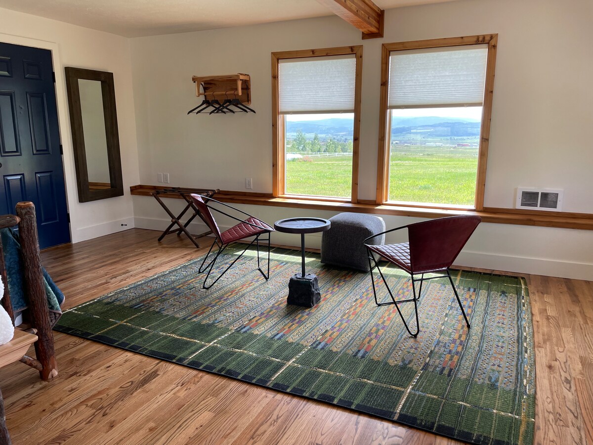 A welcoming seating area is presented with two red chairs and a low coffee table, positioned on a colorful area rug. Natural light enters through two large windows, revealing scenic views of the surrounding landscape. A wall-mounted coat rack and a wooden bench add functionality.