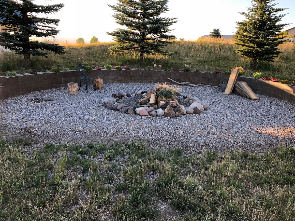 A circular fire pit area is surrounded by gravel and natural stone. Seating options include log stools and metal chairs, nestled among sparse greenery and low shrubs. Tall evergreen trees frame the space, creating a serene outdoor setting for relaxation.