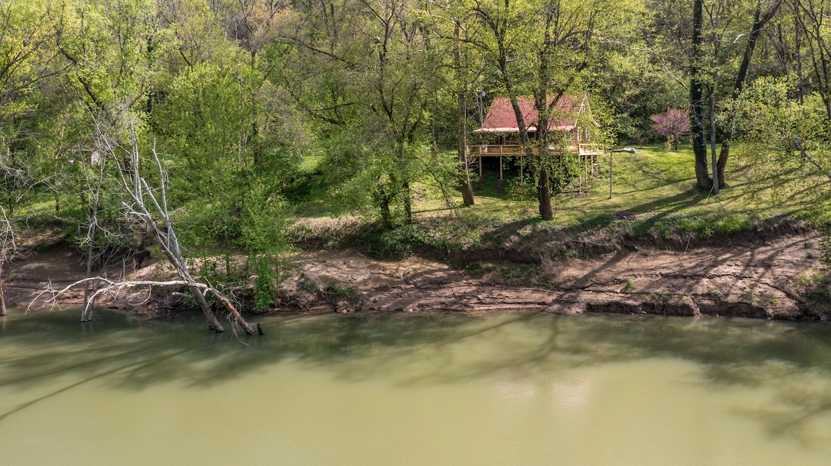 A view of the cabin is showcased amidst lush greenery, situated on a gentle slope near the riverbank. The calm water reflects the surrounding trees, and the cabin’s porch is visible, offering a serene outdoor setting.