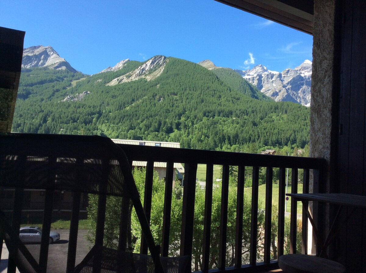 A balcony view reveals expansive green landscapes framed by mountains. Sunlight illuminates the scene, highlighting the vibrant foliage. The mountainous backdrop includes rocky peaks that rise against a clear blue sky, providing a serene escape into nature.