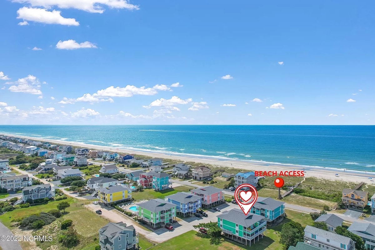 Aerial view of a coastal neighborhood highlights the proximity to the beach with clear blue waters and a sandy shore. Colorful beach houses are visible, along with designated beach access marked on the image. The sky is bright with scattered clouds.