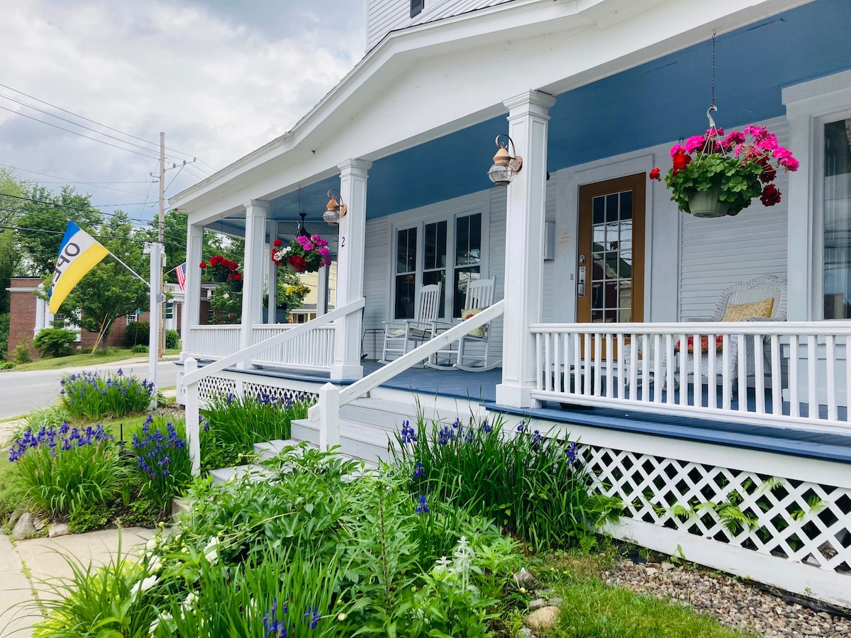 A welcoming porch is adorned with hanging flower baskets and rocking chairs, providing a relaxing outdoor space. Lush greenery and vibrant flowers line the pathway, contributing to the inviting character of the classic New England Inn's exterior.