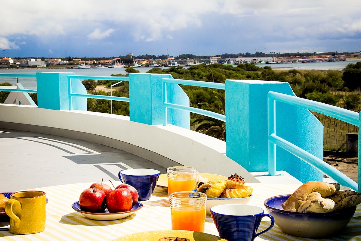 A dining table is set on a balcony with a view of the water and surrounding landscape. Fresh fruits, pastries, and beverages are displayed, with bright blue railings providing a lively contrast against the sky. The scene suggests a welcoming space for enjoying meals outdoors.