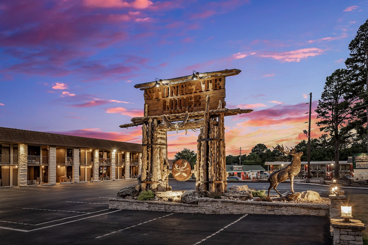 The lodge exterior is highlighted by a rustic entrance sign crafted from wood, featuring antler motifs. A spacious parking lot is visible in front, with the two-story building set against a vibrant pink and purple sunset sky, creating a tranquil atmosphere.