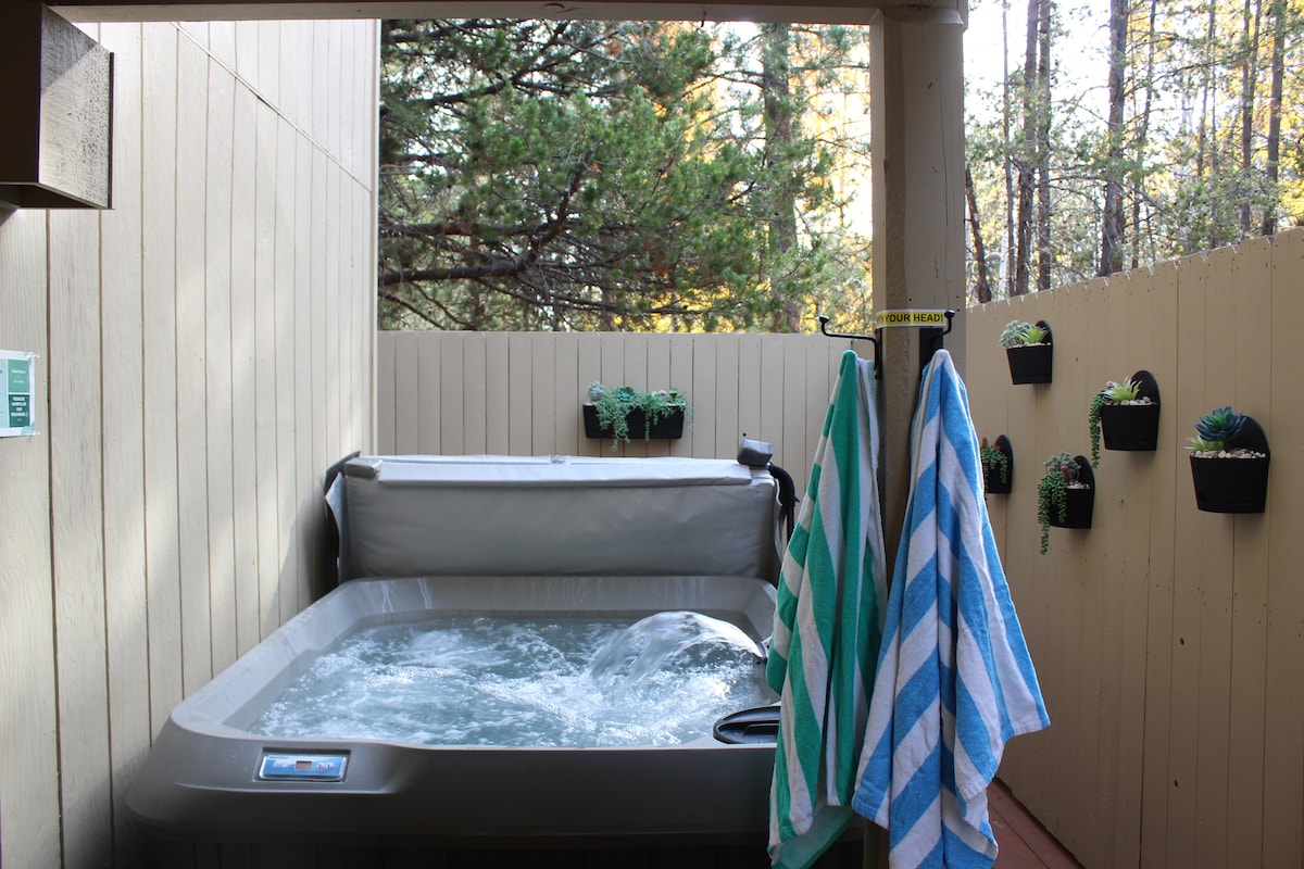 A private hot tub bubbles gently in a secluded outdoor space, surrounded by wood-paneled walls and evergreen trees. Two towels hang on a nearby railing, and small potted plants adorn the fence, adding a touch of greenery to the serene setting.
