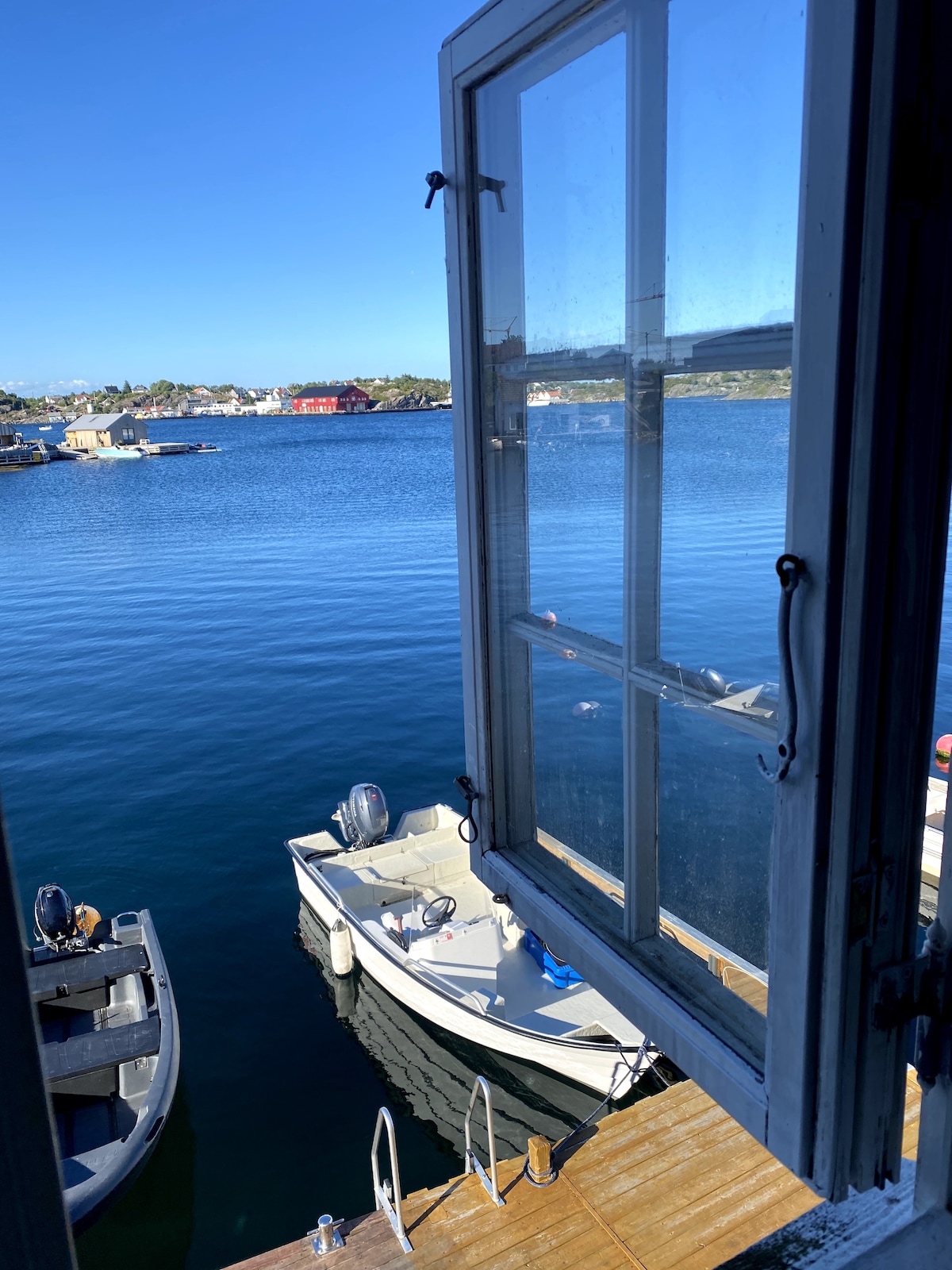 A window is opened to reveal a view of calm waters, where boats are moored along a wooden dock. Blue skies and greenery outline the serene setting, allowing natural light to illuminate the indoor space.