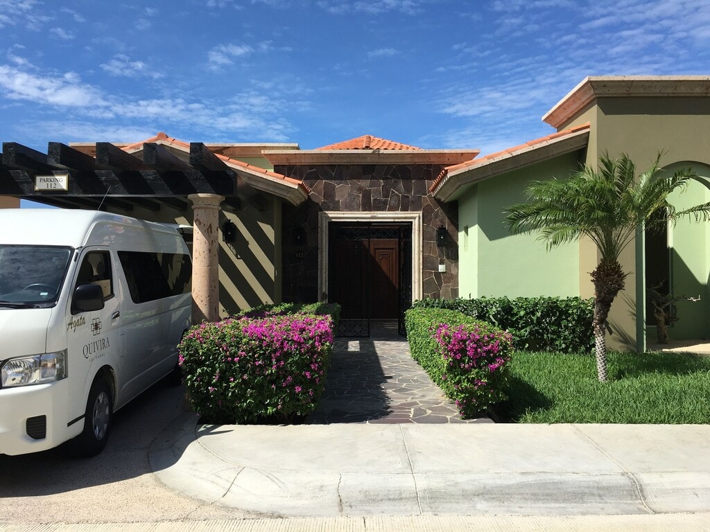 The exterior of a stylish property is showcased, featuring a combination of stone and vibrant colored walls. A pathway leads to the entrance, bordered by lush greenery and flowering shrubs. A white van is parked nearby, indicating convenient transportation options.