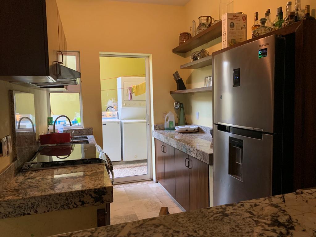 A functional kitchen is presented, featuring granite countertops and dark wooden cabinetry. Stainless steel appliances, including a refrigerator and stove, are visible. Shelves display glassware and kitchen essentials. A doorway leads to a laundry area, enhancing the space's utility.