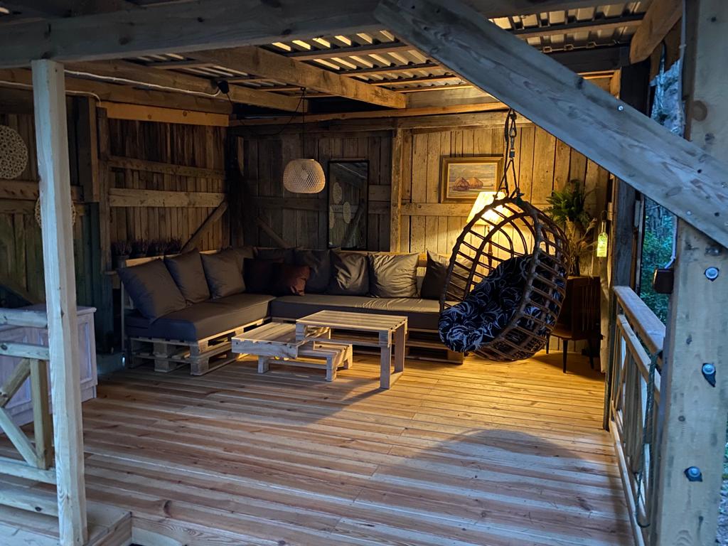 A cozy seating area is featured, showcasing a large sectional sofa with dark cushions. A round hanging chair adds a unique touch. Wooden pallets are used as low coffee tables. Soft lighting complements the rustic wooden structure, creating a comfortable atmosphere for relaxation.