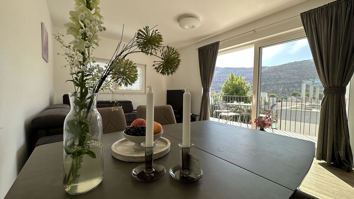 A dining table is set with two candles and a circular tray holding assorted fruits. A vase with flowers adds a touch of nature. Large glass doors open to a balcony with scenic views of mountains, creating a blend of indoor and outdoor living.
