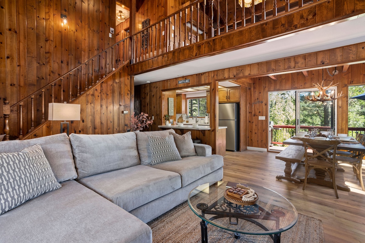 A spacious living area is highlighted by warm wood paneling and abundant natural light. A large, L-shaped sofa features neutral-toned cushions, positioned around a glass-top coffee table. In the background, a dining area and well-equipped kitchen are visible, creating an inviting communal space.