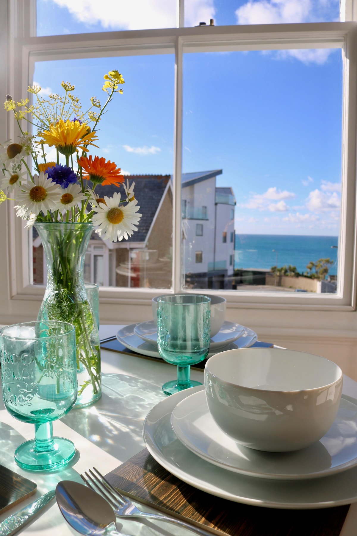 A dining table is set with white plates and aqua glasses, positioned near a window showcasing a sea view. A clear vase filled with colorful flowers adds a touch of freshness to the scene. Bright sunlight illuminates the setting, enhancing the coastal ambiance.