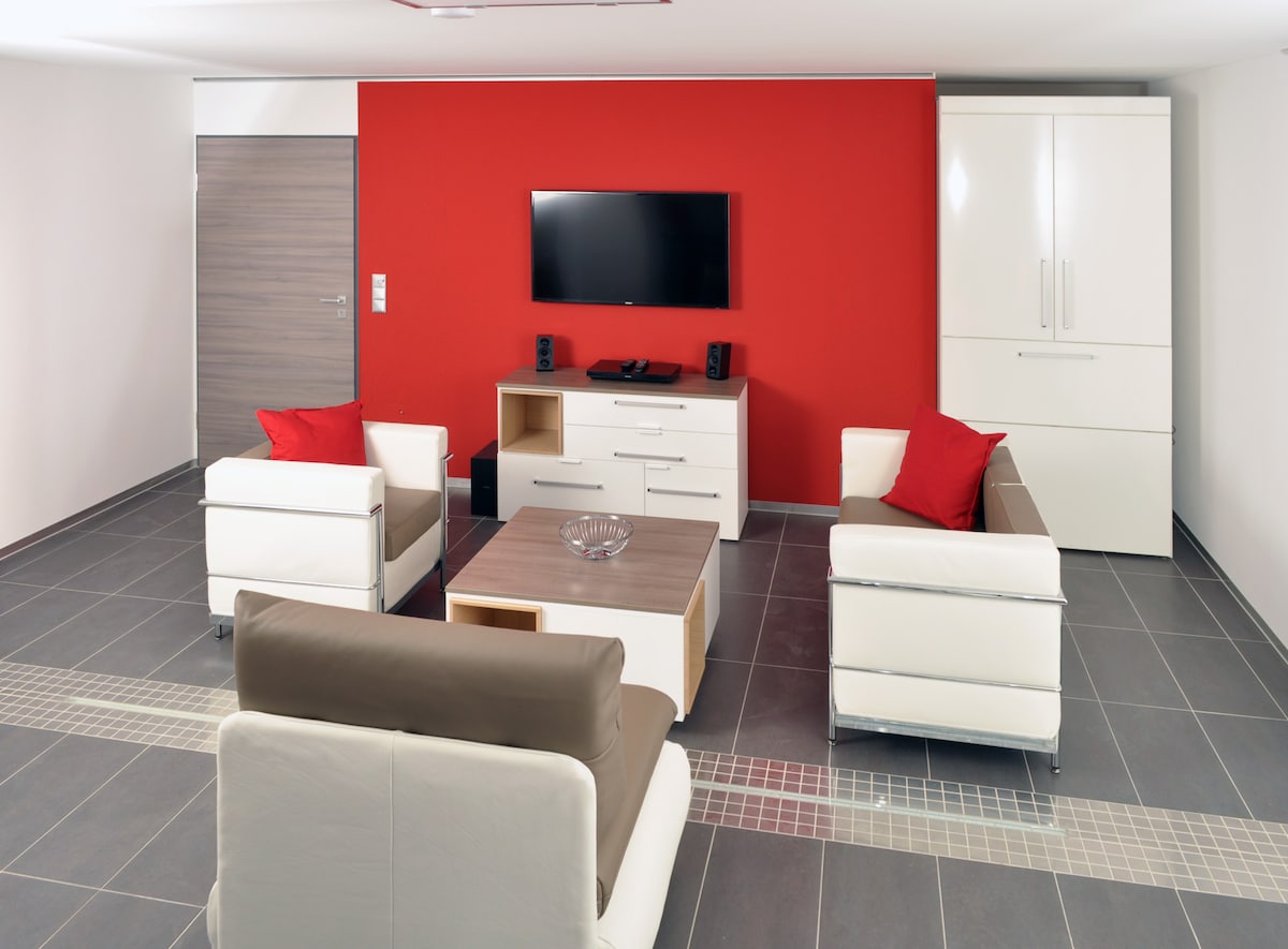 A modern living area features a red accent wall and sleek furniture. Two white armchairs with red cushions and a brown sofa surround a minimalist coffee table. A flat-screen TV is mounted on the wall, complemented by a light-colored media console.