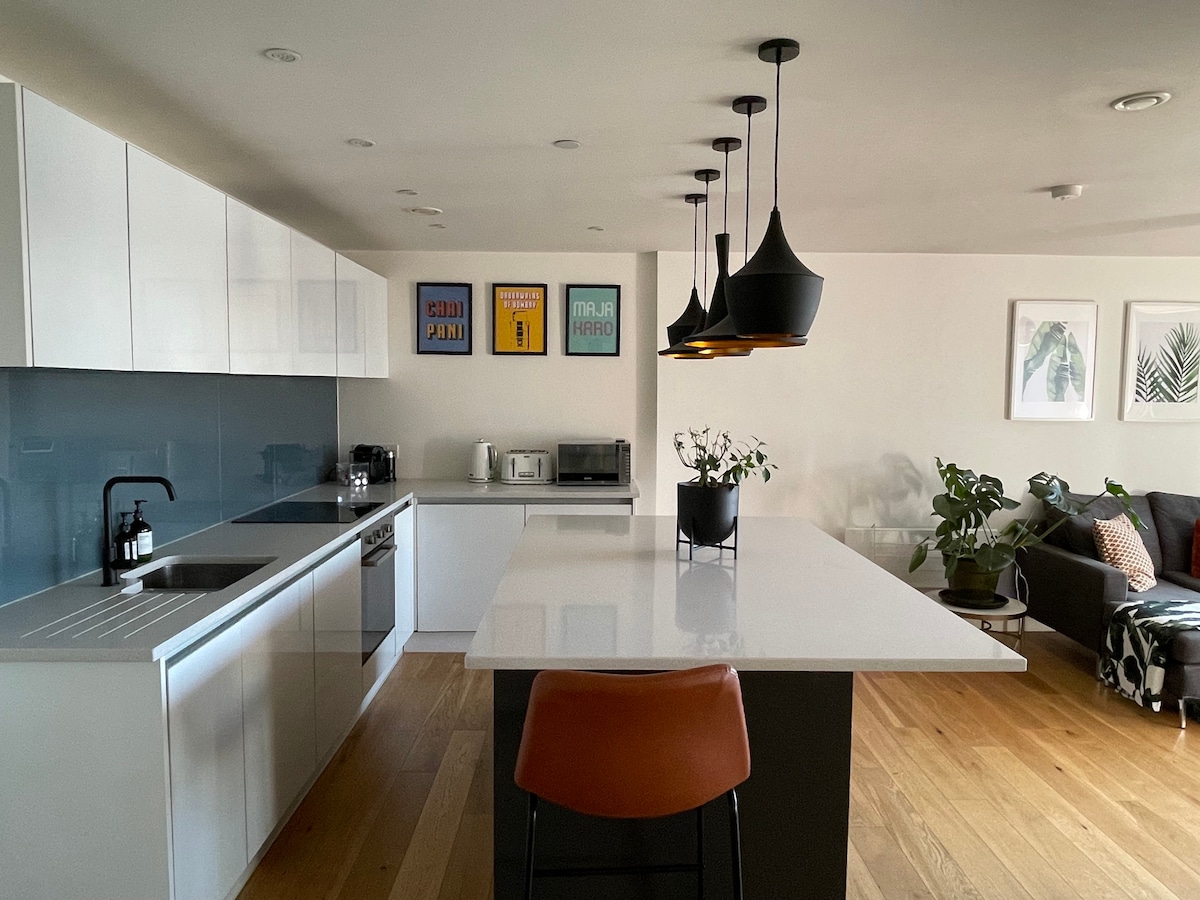 A modern kitchen features white cabinetry and a large central island with seating. Distinctive black pendant lights hang above, and artwork adorns the walls. Potted plants add a touch of greenery, while a comfortable couch is visible in the adjoining living area.