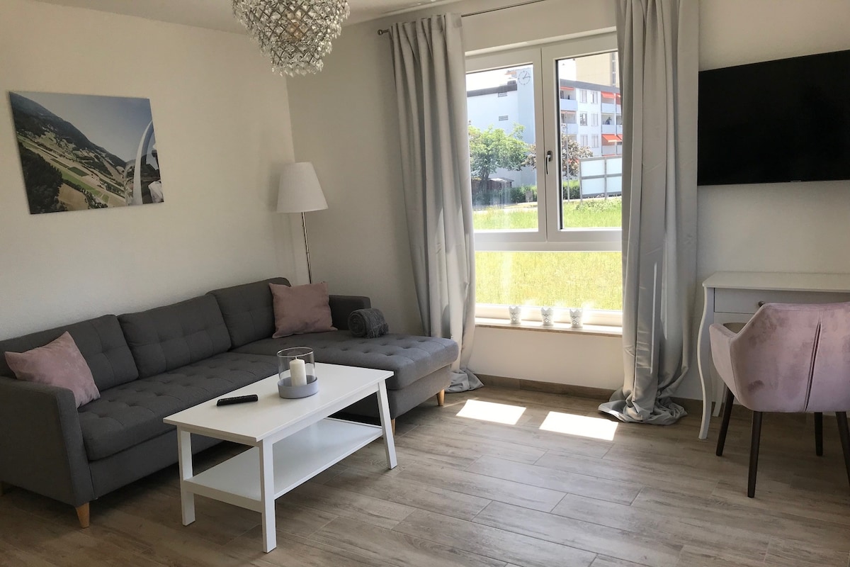 A spacious living area features a modern gray sectional sofa adorned with soft pillows. A light-colored coffee table is placed in front, with a decorative candle. Natural light flows through a large window, complemented by sheer curtains, enhancing the inviting atmosphere of the room.