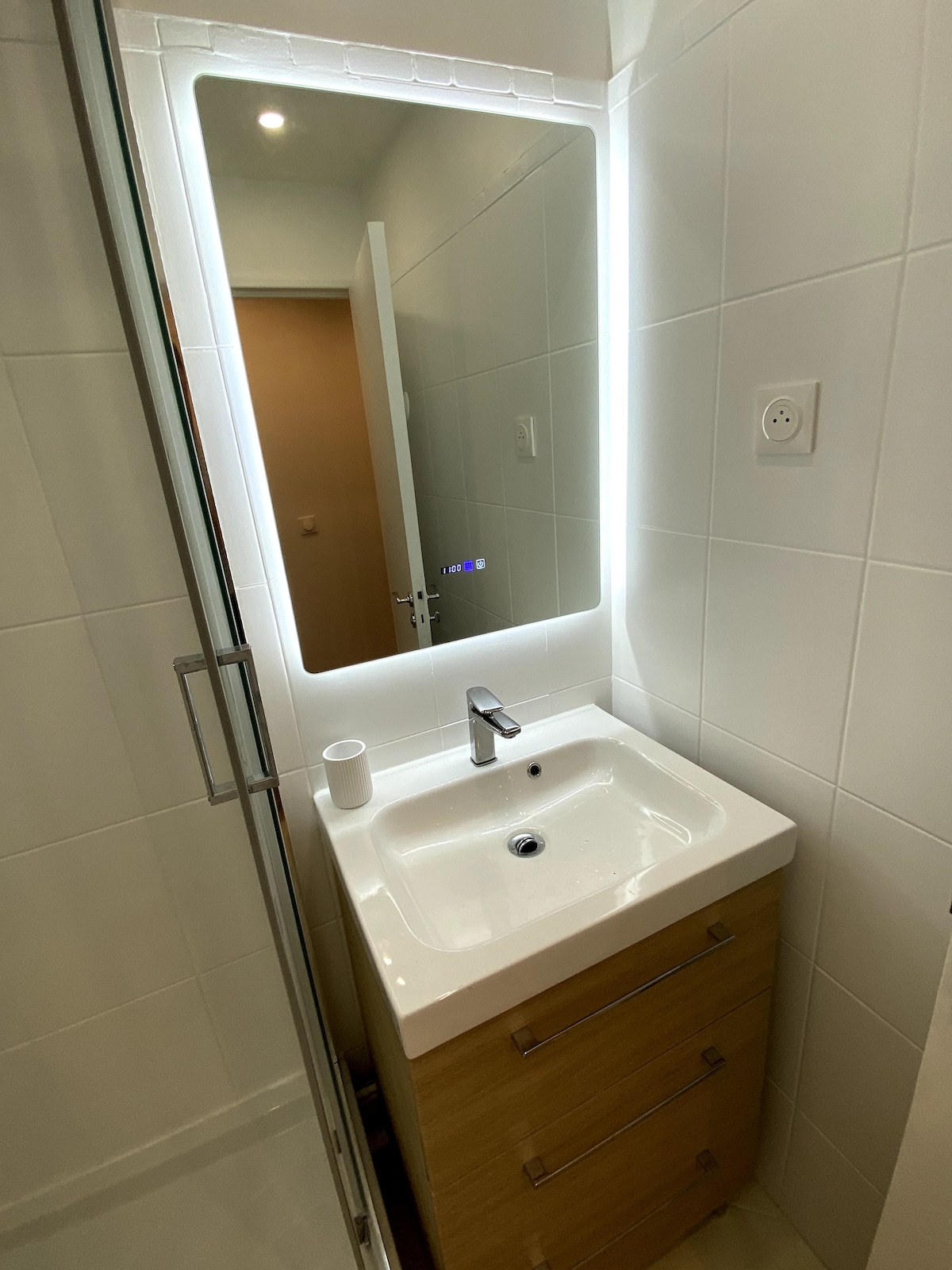 A modern bathroom is displayed, featuring a sleek vanity with a spacious sink. Above the sink, a backlit mirror enhances the area with soft illumination. Neutral tiles cover the walls, contributing to an airy and clean appearance. A glimpse of the doorway can be seen in the background.