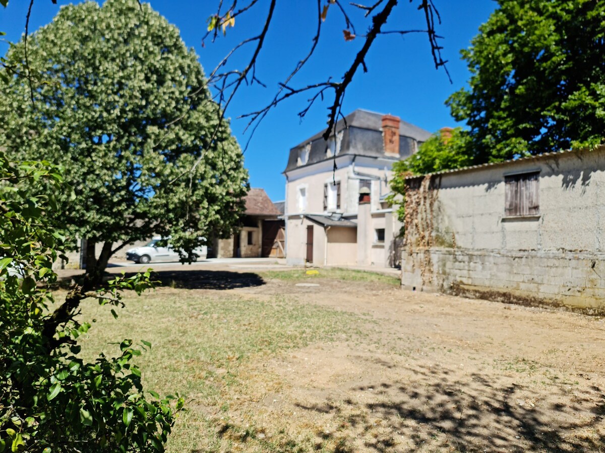 A spacious outdoor area is visible, featuring a well-maintained lawn surrounded by mature trees. A two-story building stands in the background, with a garage and an additional structure nearby. The clear blue sky adds to the open feel of the area.