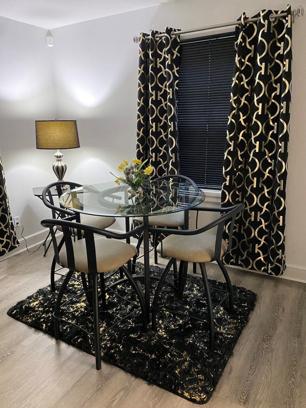 A stylish dining area features a glass-top table surrounded by four black metal chairs. A decorative rug adds texture underneath, while two patterned curtains frame a window. A floor lamp with a soft glow complements the modern decor.