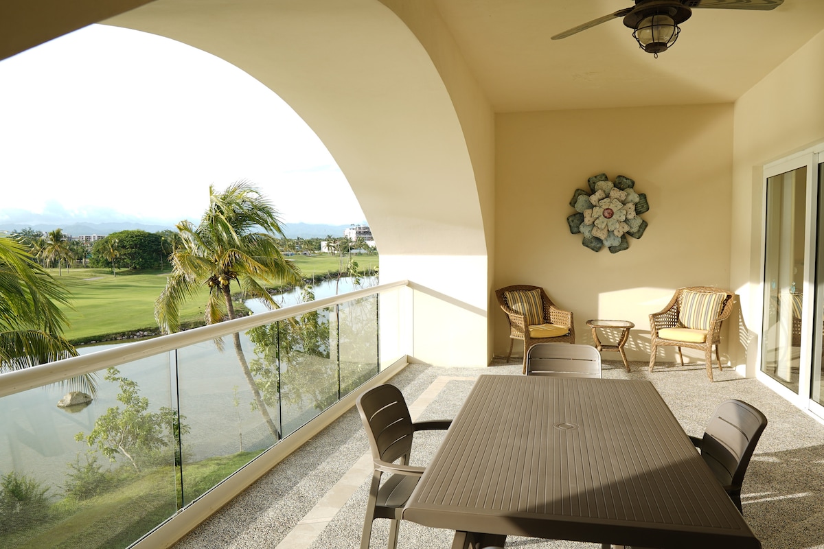 A spacious terrace offers a dining table surrounded by chairs, overlooking a lush landscape with golf course views. Two comfortable armchairs are positioned nearby, and a decorative wall piece adds visual interest to the neutral-toned wall. Natural light fills the area, creating an open and airy feel.