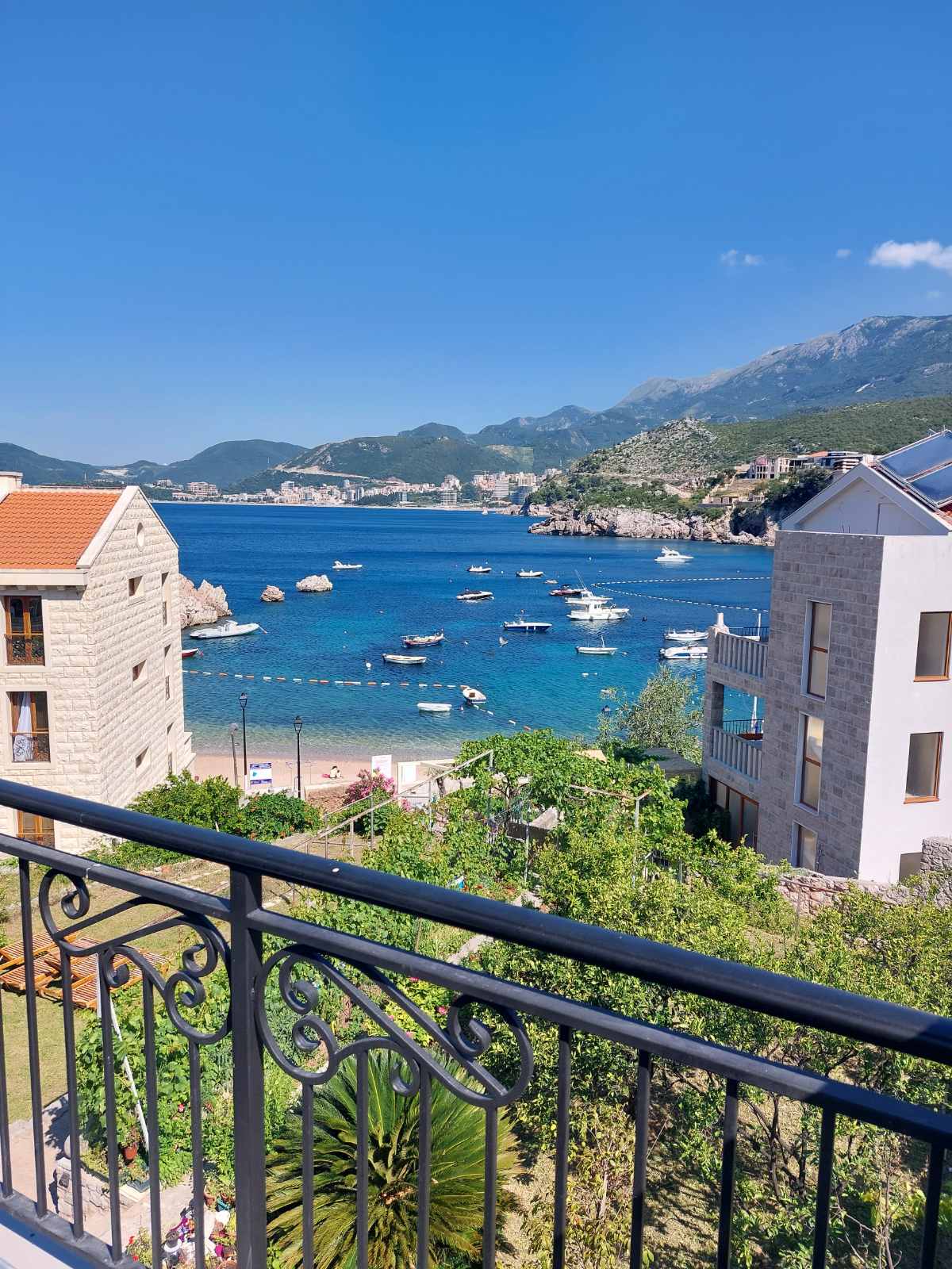 A stunning view overlooking the sea from a spacious balcony. The image captures boats gently bobbing in the clear blue water, framed by lush greenery and distant mountains. The coastline features a sandy beach, creating a serene atmosphere.