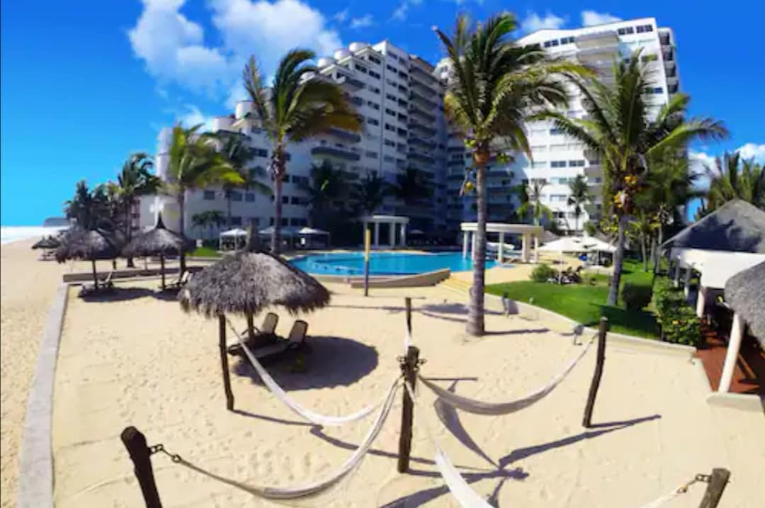 A sandy beach area is highlighted by swaying palm trees and inviting hammocks. A swimming pool is situated within a landscaped yard, surrounded by tropical greenery and lounge chairs. The building stands in the background, showcasing multiple floors and ample space for relaxation.