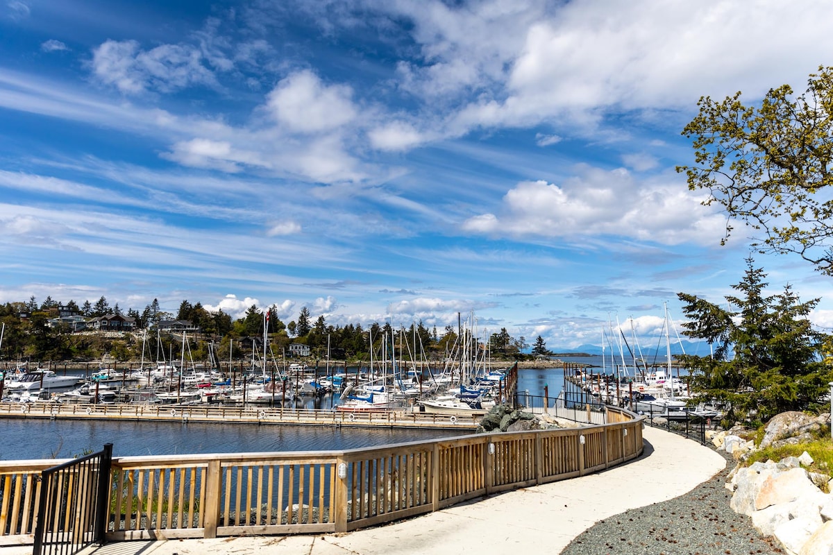 A scenic marina is visible under a bright blue sky filled with clouds. The wooden boardwalk winds along the water, leading to numerous sailboats docked in the harbor. Trees line the path, providing a natural framing to the picturesque view.