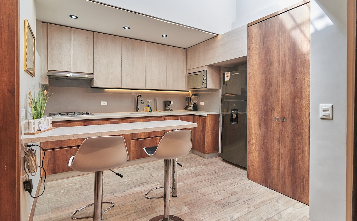 A modern kitchen is featured, equipped with wooden cabinets and granite countertops. Two bar stools are positioned at a light-colored counter. Essential appliances such as a microwave, coffee maker, and refrigerator are visible, enhancing functionality. Natural light floods the space from overhead fixtures.